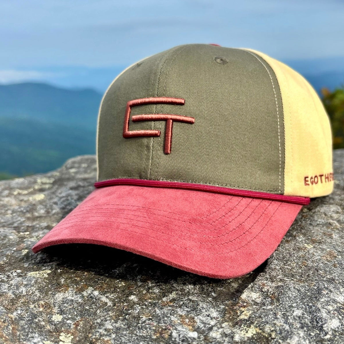 A six-panel trucker-style cap resting on a large rock with a mountain landscape and cloudy sky in the background. The cap has an olive green front panel, a pale yellow side panel, and a muted red brim, with red embroidered lettering on the front and side.